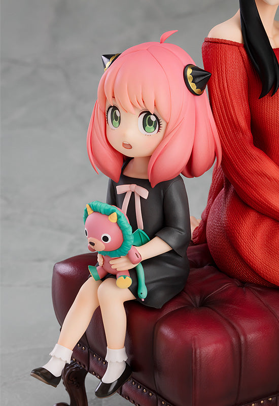 Close-up detail of Anya from GSC SPY x FAMILY 1/7 scale figure with Yor, Anya's cat ears and green eyes in pink/red outfit holding Chimera plushie on red ottoman